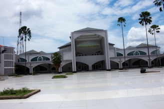 A large, modern building with a symmetrical design featuring arches and green-tinted windows. Tall palm trees are evenly spaced around the open courtyard, which has a reflective tiled surface. A cloudy sky and a communications tower are visible in the background.