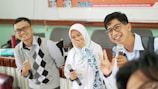 Three people are smiling and posing for a camera, holding microphones. The person in the middle is wearing a hijab and laughs with a hand covering their mouth, while the two men flanking them wear glasses and are gesturing in a friendly manner. There is a blurred foreground of another person partially visible.