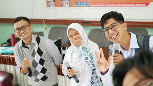Three people are smiling and posing for a camera, holding microphones. The person in the middle is wearing a hijab and laughs with a hand covering their mouth, while the two men flanking them wear glasses and are gesturing in a friendly manner. There is a blurred foreground of another person partially visible.