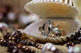 A large faceted gemstone ring is displayed inside an opened seashell. The background is adorned with various pearls and beads, scattered on a sandy surface, creating a luxurious and elegant setting.