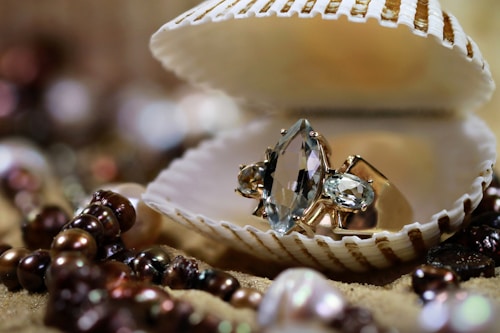 A large faceted gemstone ring is displayed inside an opened seashell. The background is adorned with various pearls and beads, scattered on a sandy surface, creating a luxurious and elegant setting.