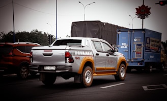 A pickup truck with a silver and yellow color scheme is prominently featured on a road. It is surrounded by other vehicles, including an orange SUV and a blue delivery truck. The background includes trees, road signs, and traffic lights under an overcast sky.