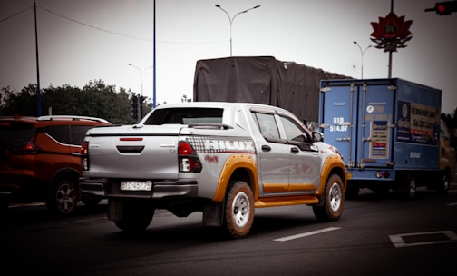 A pickup truck with a silver and yellow color scheme is prominently featured on a road. It is surrounded by other vehicles, including an orange SUV and a blue delivery truck. The background includes trees, road signs, and traffic lights under an overcast sky.