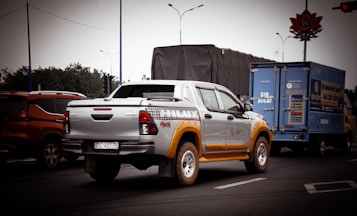A pickup truck with a silver and yellow color scheme is prominently featured on a road. It is surrounded by other vehicles, including an orange SUV and a blue delivery truck. The background includes trees, road signs, and traffic lights under an overcast sky.