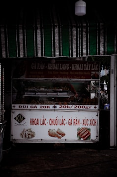 A small street food stall with signage advertising various fried foods such as sweet potatoes, bananas, fried chicken, and sausages. The stall has colorful images of these foods and displays pricing details. It is located under a green striped canopy with a visible light fixture above.
