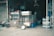 A cluttered storefront displays various goods including animal cages, a broom, a scale, and several containers. The entrance is dark with shelves and stacked items visible in the background.