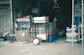 A cluttered storefront displays various goods including animal cages, a broom, a scale, and several containers. The entrance is dark with shelves and stacked items visible in the background.