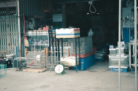 A cluttered storefront displays various goods including animal cages, a broom, a scale, and several containers. The entrance is dark with shelves and stacked items visible in the background.