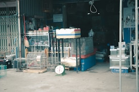 A cluttered storefront displays various goods including animal cages, a broom, a scale, and several containers. The entrance is dark with shelves and stacked items visible in the background.