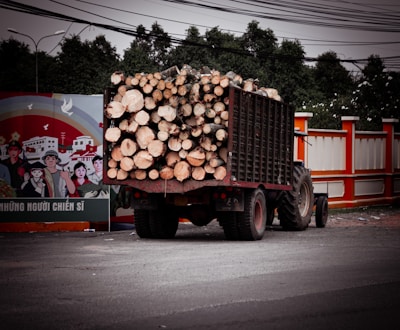 A tractor is loaded with a large stack of cut logs, parked near a colorful mural featuring a group of people and buildings. The background includes trees and power lines, adding to the rural and industrial setting.