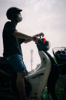 A person wearing a helmet and mask is sitting on a parked scooter. In the background, there is a tall communication tower and some greenery.