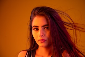 A woman with long flowing hair is featured against a vibrant orange background. Her facial expression is calm and composed, with her eyes looking directly at the viewer. The lighting casts warm hues on her face, highlighting her makeup and features.