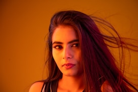 A woman with long flowing hair is featured against a vibrant orange background. Her facial expression is calm and composed, with her eyes looking directly at the viewer. The lighting casts warm hues on her face, highlighting her makeup and features.