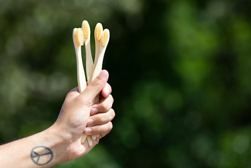 A hand holds three bamboo toothbrushes with light bristles against a blurred green natural background. A tattoo resembling a peace symbol is visible on the wrist.
