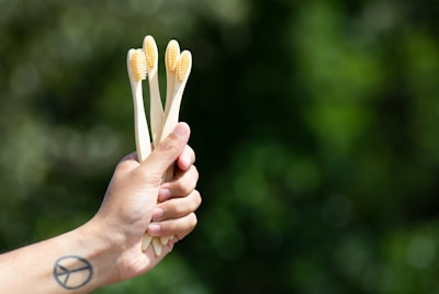 A hand holds three bamboo toothbrushes with light bristles against a blurred green natural background. A tattoo resembling a peace symbol is visible on the wrist.