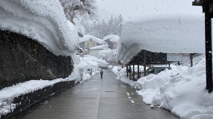 A lane in Nozawa Onsen village hemmed in by enormous snowbanks as snow falls, with a lone figure walking through