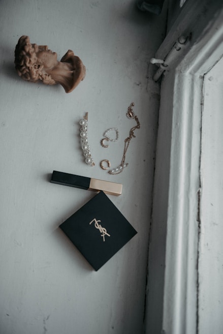 A flat lay composition features a decorative bust, a pearl hairpin, a chain necklace, a makeup compact with a logo, and a tube of lip gloss. The items are arranged on a light-colored surface next to a window, creating a serene and minimalist aesthetic.
