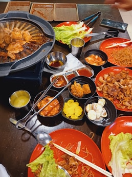 A tabletop filled with various dishes including grilled meat cooking on a hot plate, fresh lettuce, noodles, and assorted side dishes like pickled vegetables and sauces. The setting has a casual dining atmosphere with plates and utensils scattered around.