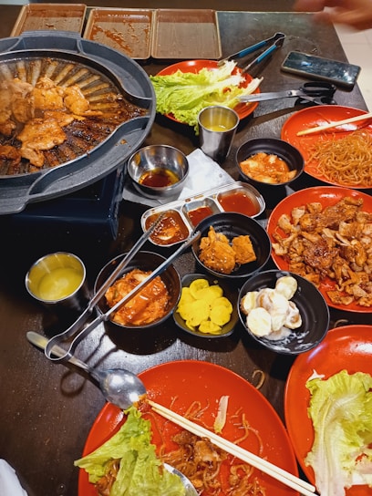 A tabletop filled with various dishes including grilled meat cooking on a hot plate, fresh lettuce, noodles, and assorted side dishes like pickled vegetables and sauces. The setting has a casual dining atmosphere with plates and utensils scattered around.