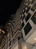 A tall, modern skyscraper with a grid-like facade of glass and steel, extending upward into a dark night sky. The building features a sign reading 'MODEL OPEN' and has illuminated windows and balconies. Another cylindrical glass building is partially visible in the background.