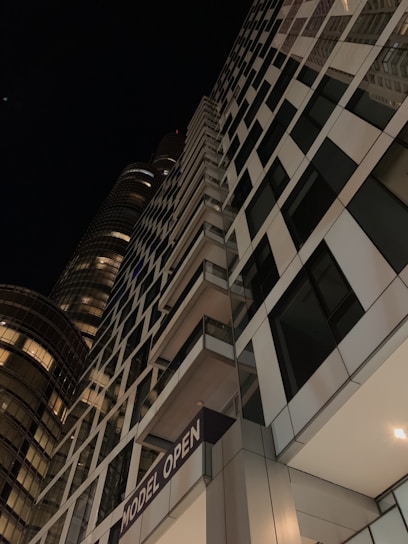 A tall, modern skyscraper with a grid-like facade of glass and steel, extending upward into a dark night sky. The building features a sign reading 'MODEL OPEN' and has illuminated windows and balconies. Another cylindrical glass building is partially visible in the background.