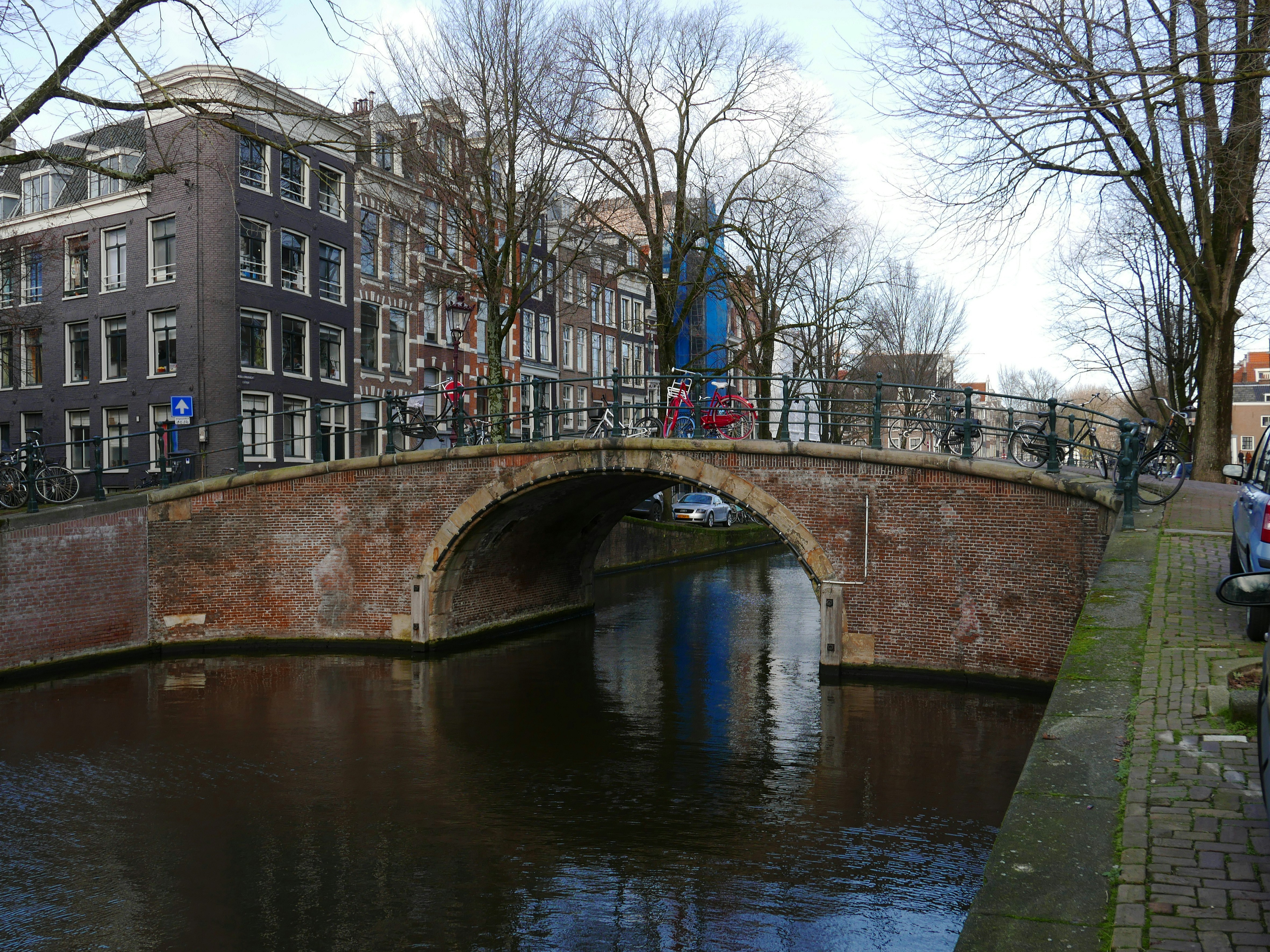 Photo of old brick bridge with one arch at the canal water Reguliersgracht with winter trees. This bridge is part of the street Kerkstraat in Amsterdam old city. The historical canal is connecting the main canals Keizersgracht and Prinsengracht. I like very much the texture of the old bricks and the reflections and shades in the canal water. Free old city photo of Amsterdam streetviews - Dutch Mokum street photography by Fons Heijnsbroek, January 2022, The Netherlands