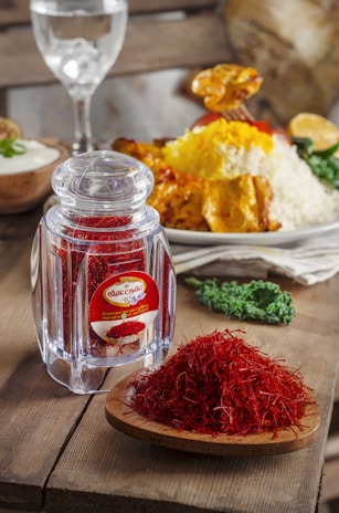 A clear glass jar filled with red saffron threads sits next to a wooden plate also holding a heap of saffron. In the background, a serving of rice garnished with yellow spices and roasted chicken is visible along with a glass of water and a bowl of yogurt. The setting appears to be a rustic kitchen or dining area.