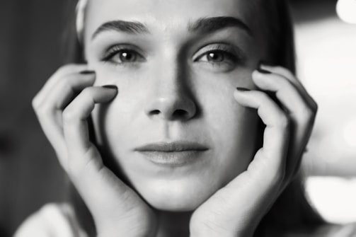 A close-up black and white portrait of a person with intense, thoughtful expression. The individual's hands gently frame their face, adding a sense of introspection. The background is softly blurred, ensuring full attention is drawn to the facial expression and details.