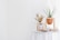 On a round white table, a wooden stand holds a terracotta pot with a tall green plant. Beside it, another stand contains dried flowers and a glass jar. The scene is serene with minimalistic decor and neutral tones, enhanced by scattered dried elements.