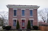A symmetrical two-story brick house with white trim and a prominent red door is flanked by tall evergreen shrubs. It features large windows with white sills and lintels. The roof is covered with red shingles, and there is a wooden fence on the right side of the house. Leafless trees surround the property, indicating a winter or early spring setting.