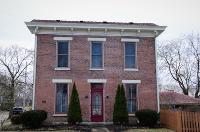 A symmetrical two-story brick house with white trim and a prominent red door is flanked by tall evergreen shrubs. It features large windows with white sills and lintels. The roof is covered with red shingles, and there is a wooden fence on the right side of the house. Leafless trees surround the property, indicating a winter or early spring setting.