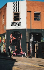 An urban street corner features a large mural of a person's profile with a glowing neon effect around the neck. The wall is part of a building made of red brick and white paint. Above the mural, the words 'London Shuffle Club' are written. A person is walking by, carrying a shopping bag, while a bicycle is parked against the wall. The scene captures the essence of vibrant street art.