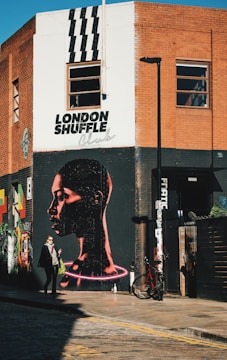 An urban street corner features a large mural of a person's profile with a glowing neon effect around the neck. The wall is part of a building made of red brick and white paint. Above the mural, the words 'London Shuffle Club' are written. A person is walking by, carrying a shopping bag, while a bicycle is parked against the wall. The scene captures the essence of vibrant street art.
