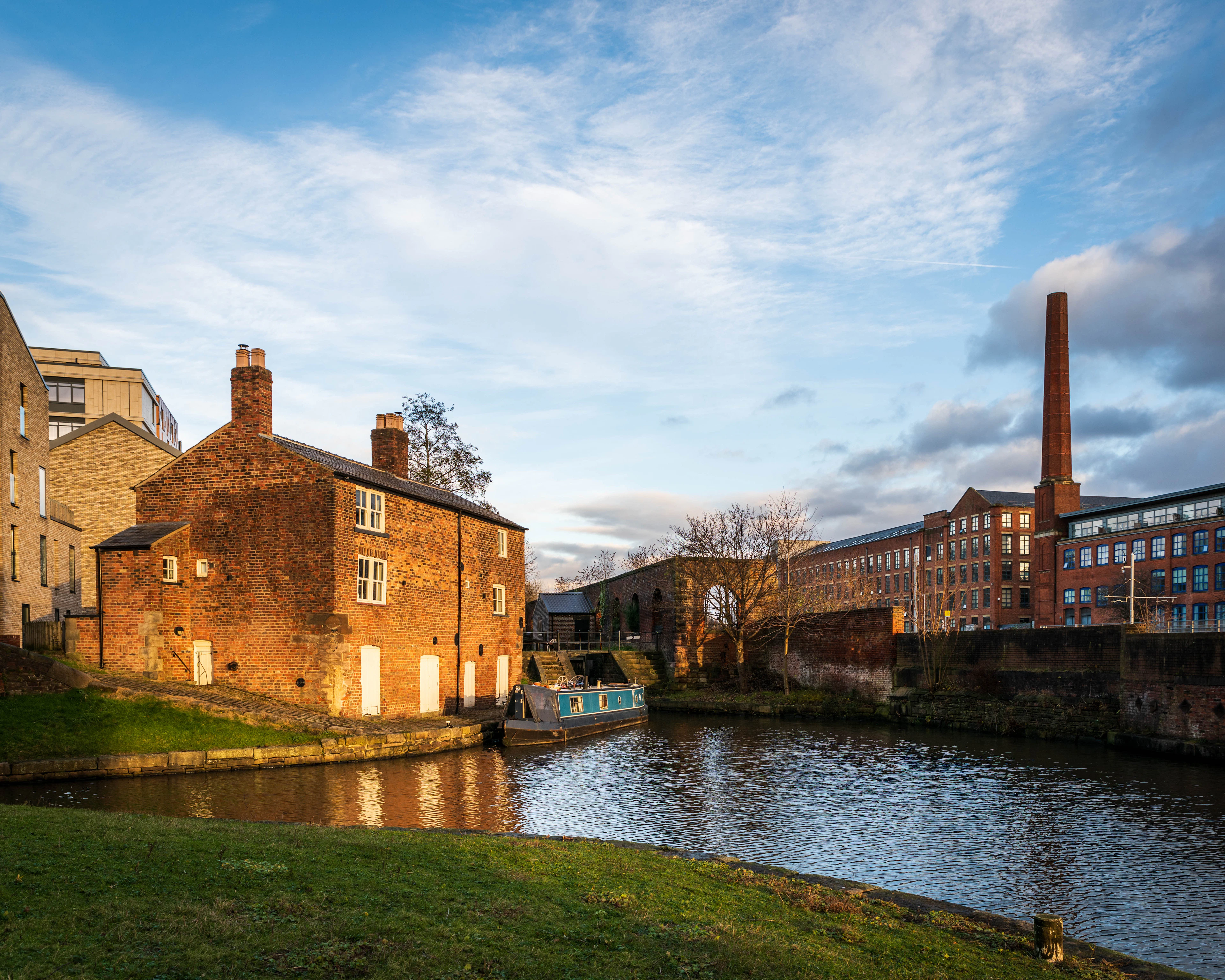 The old lock keeper's cottage on the canal at Castlefield in Manchester in low golden light, a moored narrowboat beside it and a red-brick mill and chimney across the still water