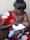 A woman in a red floral dress is breastfeeding a baby, tenderly holding the child in her arms while sitting on a couch. The scene is intimate and nurturing, showcasing a close bond between mother and child.