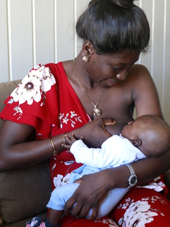 A woman in a red floral dress is breastfeeding a baby, tenderly holding the child in her arms while sitting on a couch. The scene is intimate and nurturing, showcasing a close bond between mother and child.