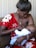 A woman in a red floral dress is breastfeeding a baby, tenderly holding the child in her arms while sitting on a couch. The scene is intimate and nurturing, showcasing a close bond between mother and child.