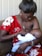 A woman in a red floral dress is breastfeeding a baby, tenderly holding the child in her arms while sitting on a couch. The scene is intimate and nurturing, showcasing a close bond between mother and child.