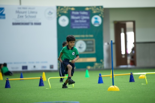 A young boy in a green shirt and dark shorts is actively participating in an indoor obstacle course. The room features green flooring and various colorful cones and poles arranged to form hurdles. Background elements include banners and signs, indicating a school setting.