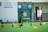A young boy in a green shirt and dark shorts is actively participating in an indoor obstacle course. The room features green flooring and various colorful cones and poles arranged to form hurdles. Background elements include banners and signs, indicating a school setting.