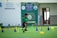 A young boy in a green shirt and dark shorts is actively participating in an indoor obstacle course. The room features green flooring and various colorful cones and poles arranged to form hurdles. Background elements include banners and signs, indicating a school setting.