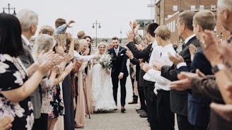 A newlywed couple walks hand-in-hand down an outdoor aisle as guests line both sides clapping and cheering. The bride is dressed in a flowing white gown holding a bouquet, while the groom wears a dark suit. The guests include people of various ages, dressed in formal attire, creating an atmosphere of celebration and happiness.