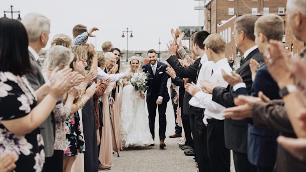 A newlywed couple walks hand-in-hand down an outdoor aisle as guests line both sides clapping and cheering. The bride is dressed in a flowing white gown holding a bouquet, while the groom wears a dark suit. The guests include people of various ages, dressed in formal attire, creating an atmosphere of celebration and happiness.