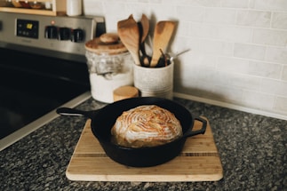 A loaf of artisan bread sits in a black cast-iron skillet on a wooden cutting board. The bread has a beautifully scored crust, highlighting a golden brown color. Nearby, a jar of flour and a collection of wooden utensils are arranged on the countertop, which is made of dark stone.