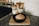 A loaf of artisan bread sits in a black cast-iron skillet on a wooden cutting board. The bread has a beautifully scored crust, highlighting a golden brown color. Nearby, a jar of flour and a collection of wooden utensils are arranged on the countertop, which is made of dark stone.