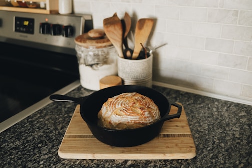 A loaf of artisan bread sits in a black cast-iron skillet on a wooden cutting board. The bread has a beautifully scored crust, highlighting a golden brown color. Nearby, a jar of flour and a collection of wooden utensils are arranged on the countertop, which is made of dark stone.