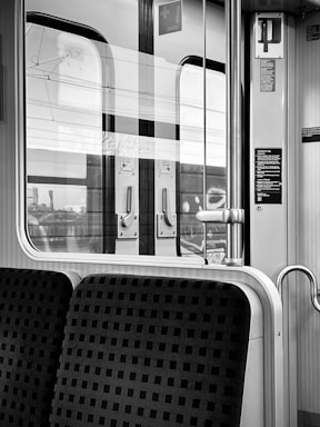 The interior of a train features patterned seats next to a window, with reflections on the glass showing parts of the outside environment. The door includes informational text and a handle for opening. Metal and plastic elements are prominent in the minimalist design.
