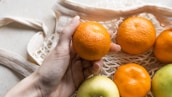 A hand is holding an orange while other oranges and a green apple rest in a reusable mesh bag. The scene suggests an eco-friendly approach to carrying fresh fruit.