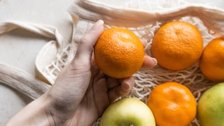 A hand is holding an orange while other oranges and a green apple rest in a reusable mesh bag. The scene suggests an eco-friendly approach to carrying fresh fruit.