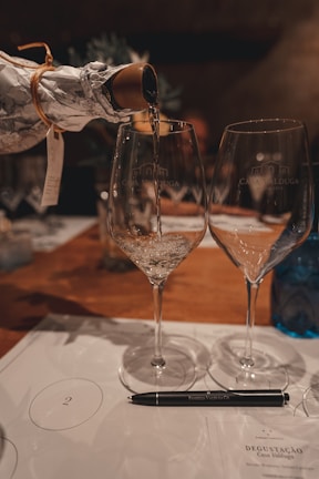 A bottle wrapped in paper is pouring a clear liquid into two elegant wine glasses with 'Casa Valduga' embossed on them. The setting includes a wooden table surface with a pen placed on a paper labeled 'Degustação'. The background is softly blurred, giving a warm and inviting atmosphere.